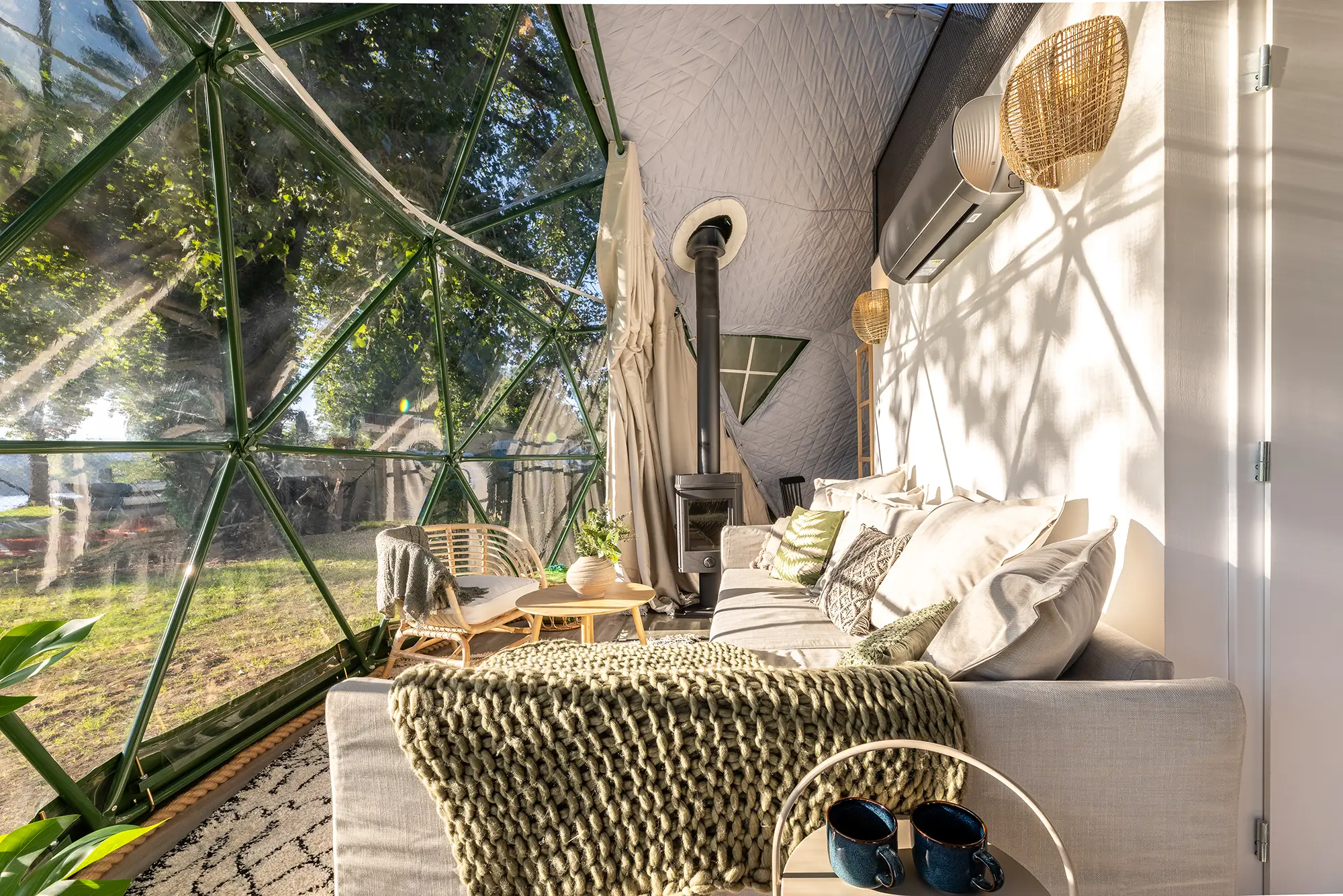 Inside a geodesic dome — sofa with throw blanket, wood-burning stove, and a view of the treeline through the glass facade